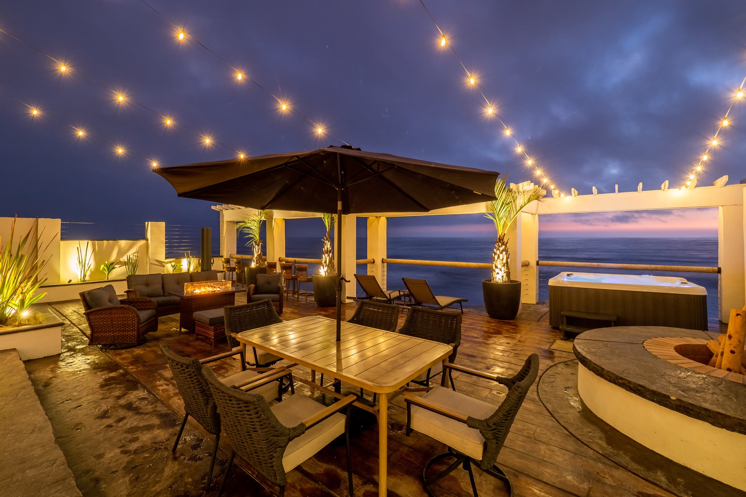 Oceanfront deck at dusk with string lights, dining, jacuzzi and fire pit